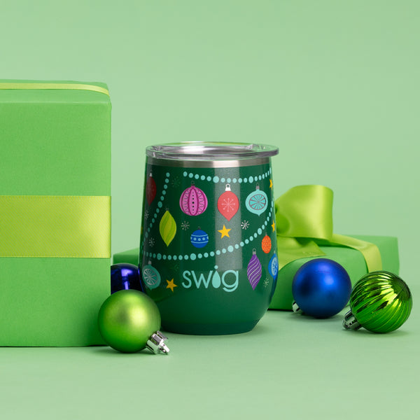 Swig Life 12oz O Christmas Tree Stemless Wine Cup on a green background next to Christmas ornaments and a green gift box