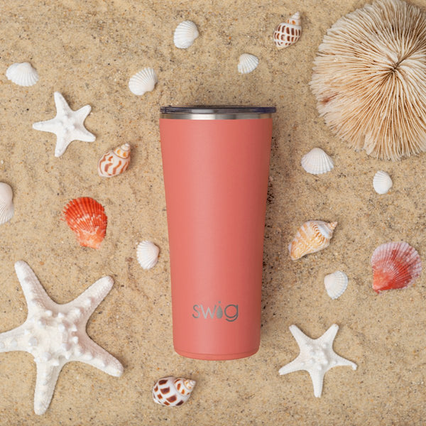 Swig Life 22oz Insulated Coral Tumbler on a sandy beach with sea shells